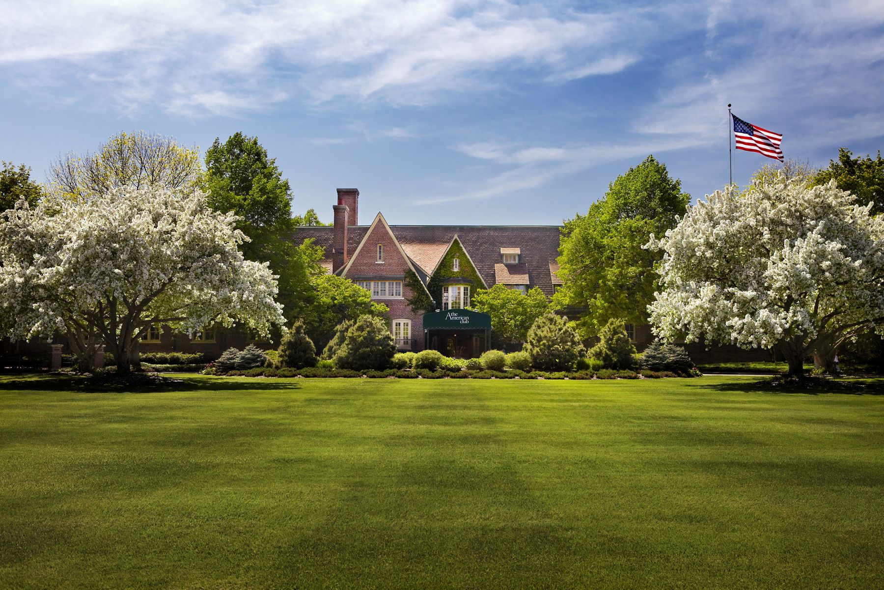 An exterior view of the brick façade and gables of the KOHLER American Club resort hotel and Kohler Waters Spa, surrounded by trees and wide manicured lawns.