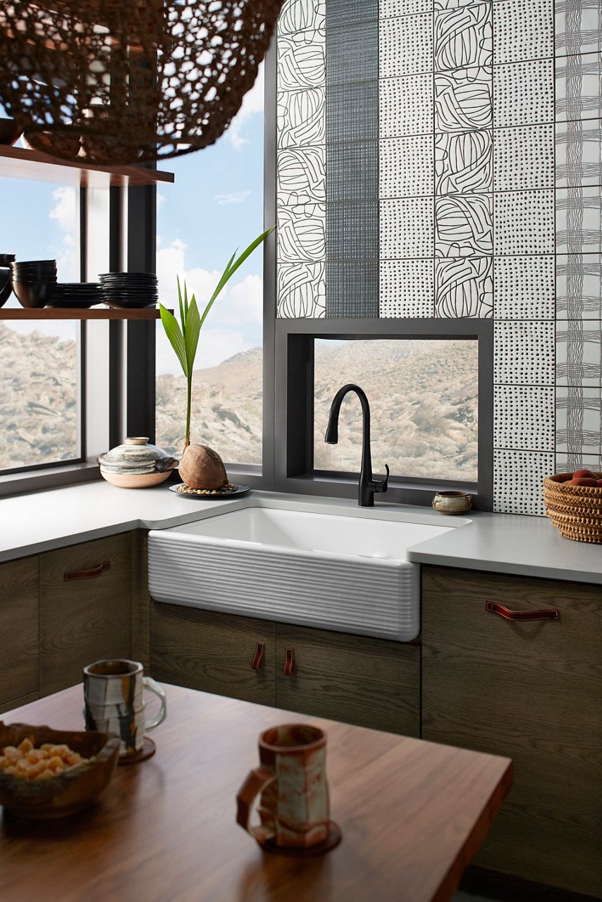 Farmhouse apron-front sink with a matte black faucet in a bright kitchen with large windows.