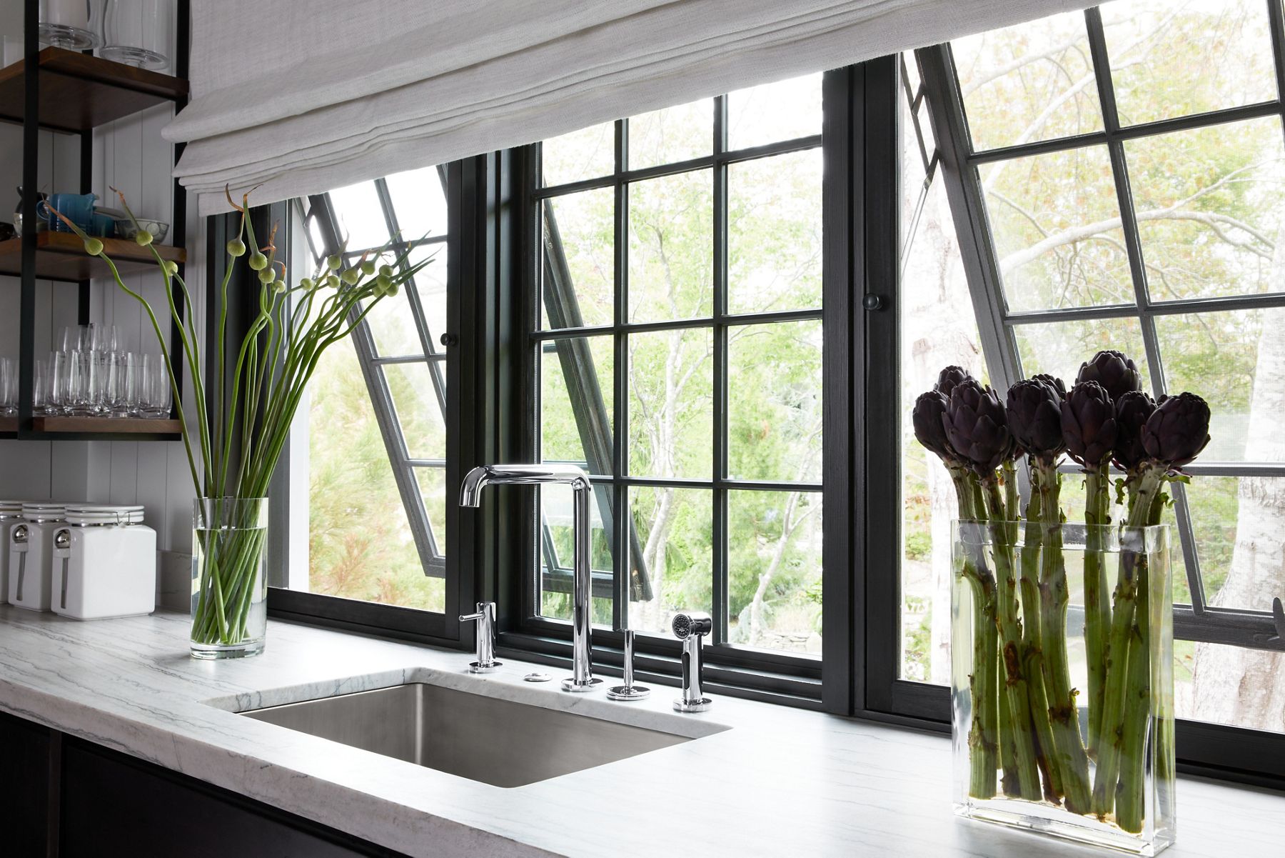 Vue sur un comptoir de cuisine en marbre avec de la verdure dans des vases en verre et des fenêtres à cadre noir qui s’inclinent pour s’ouvrir, avec des étagères ouvertes.