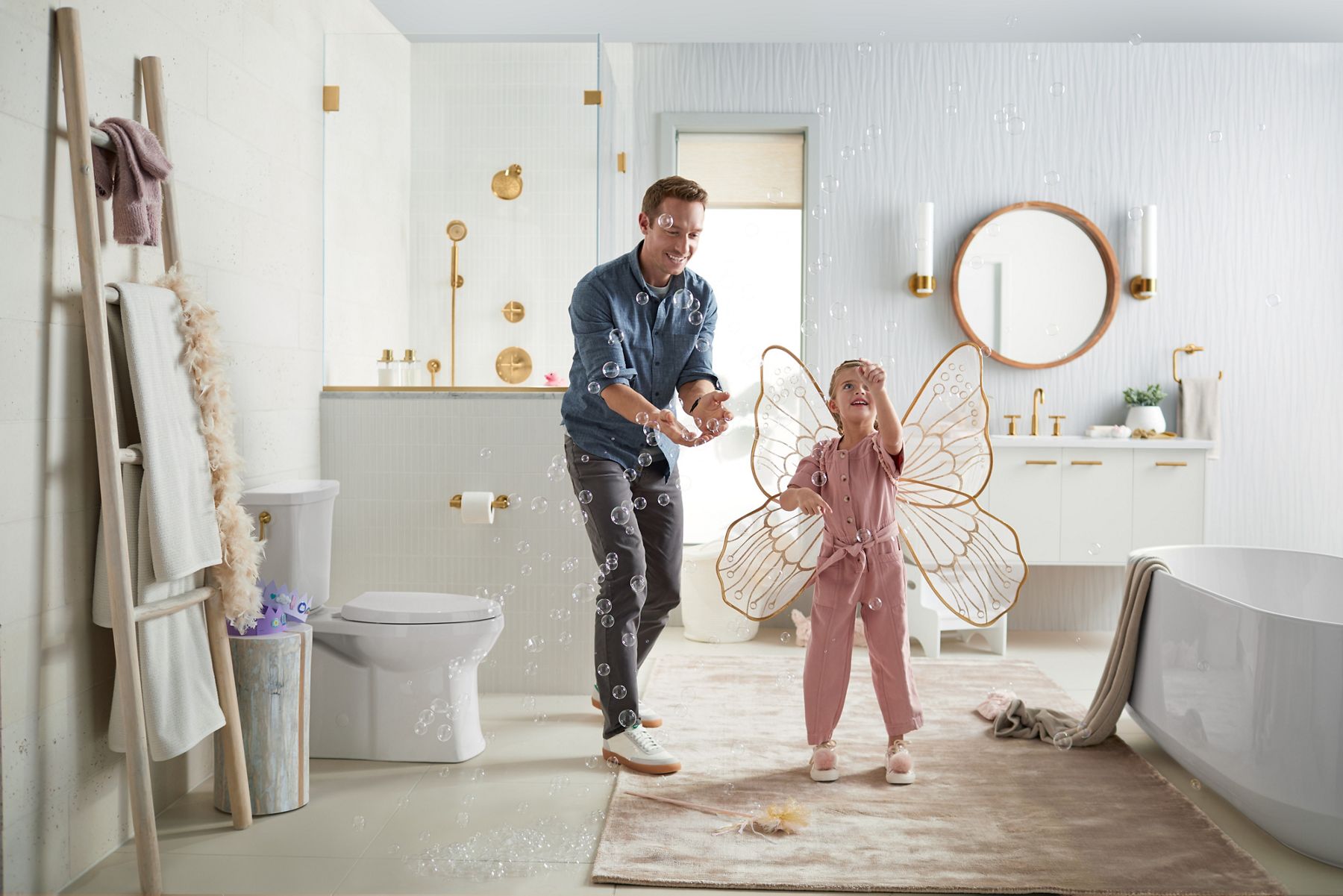 A girl wearing butterfly wings blows buttles in a white bathroom with gold accents while her father looks on and smiles.