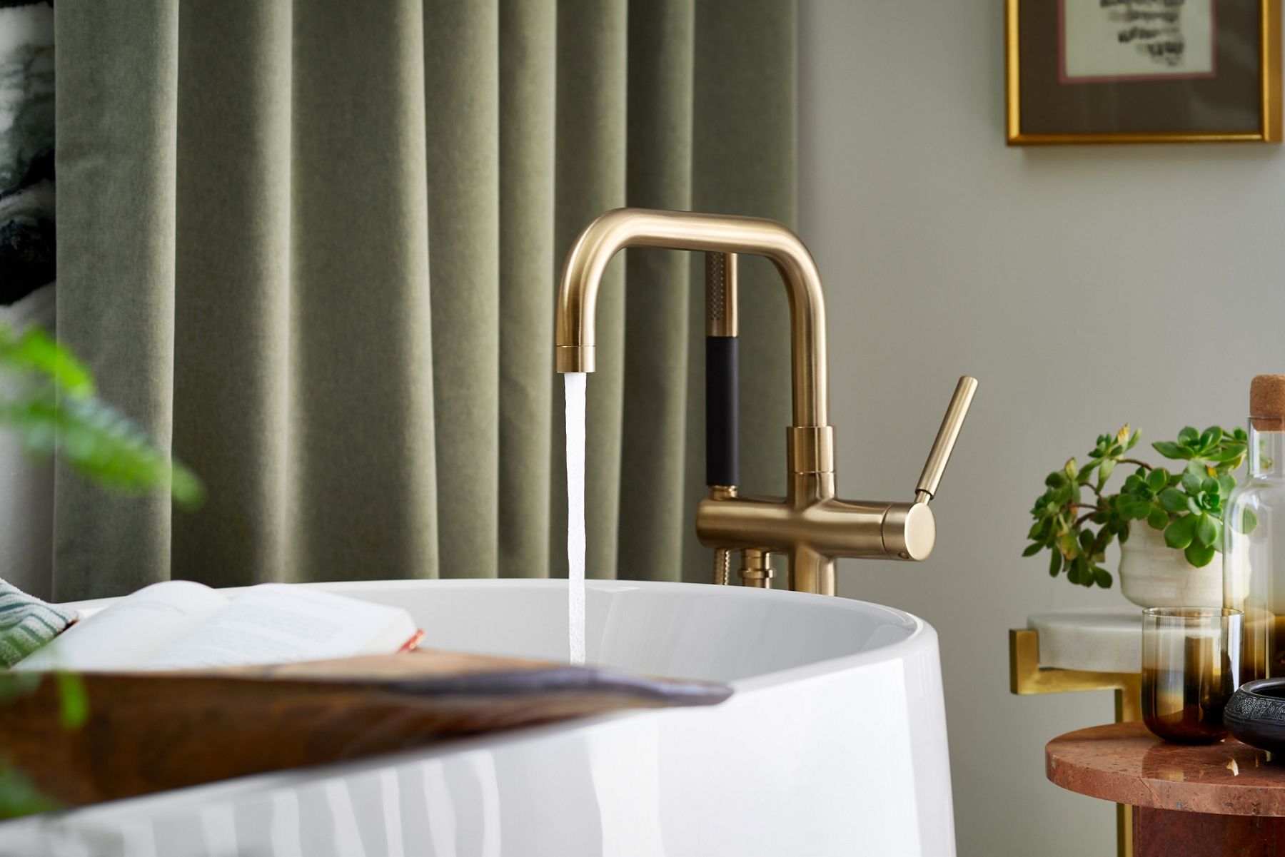 Water pouring from a brushed brass bath filling into a white freestanding bath with a tray across its top.