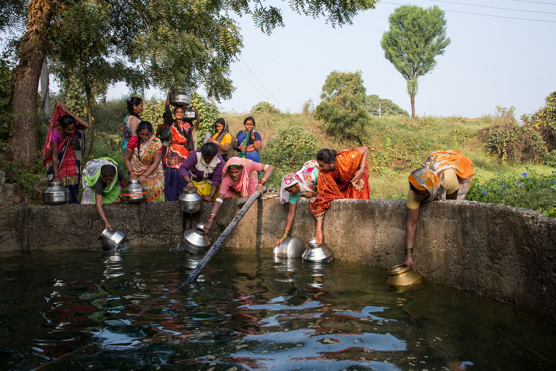 Women in colorful saris dip metal pots into an open water source.