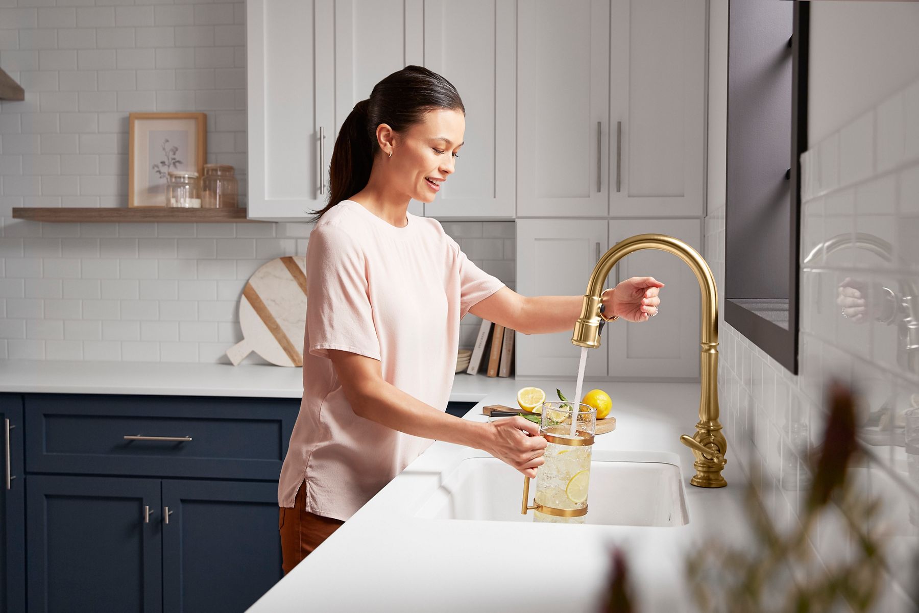 A woman in a light pink top is filling a glass pitcher with water from a stylish gold faucet in a modern kitchen. The pitcher contains slices of lemon and ice, while a cutting board with additional lemons is placed on the white countertop. The kitchen features white cabinetry and a clean, minimalist design.
