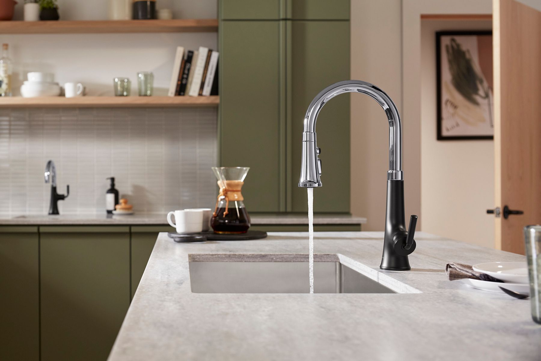 Contemporary kitchen with a sleek, modern faucet in a combination of chrome and black finishes. Water flows from the faucet into a stainless steel sink. The background features light-colored cabinetry and subtle decor, giving the space a minimalist aesthetic.
