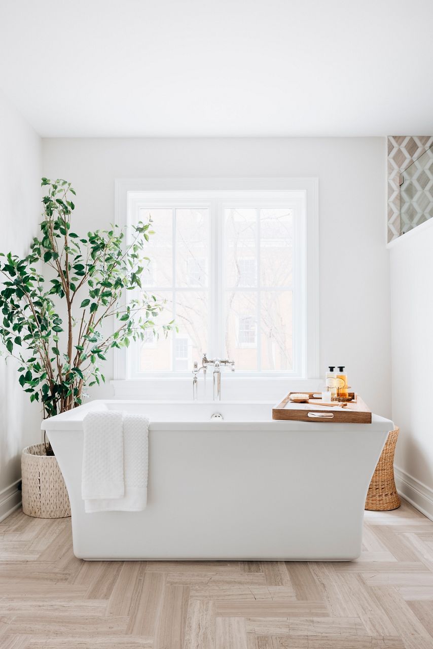 A bathroom with a white freestanding bath in an open area with a large plant and a sun-filled window.