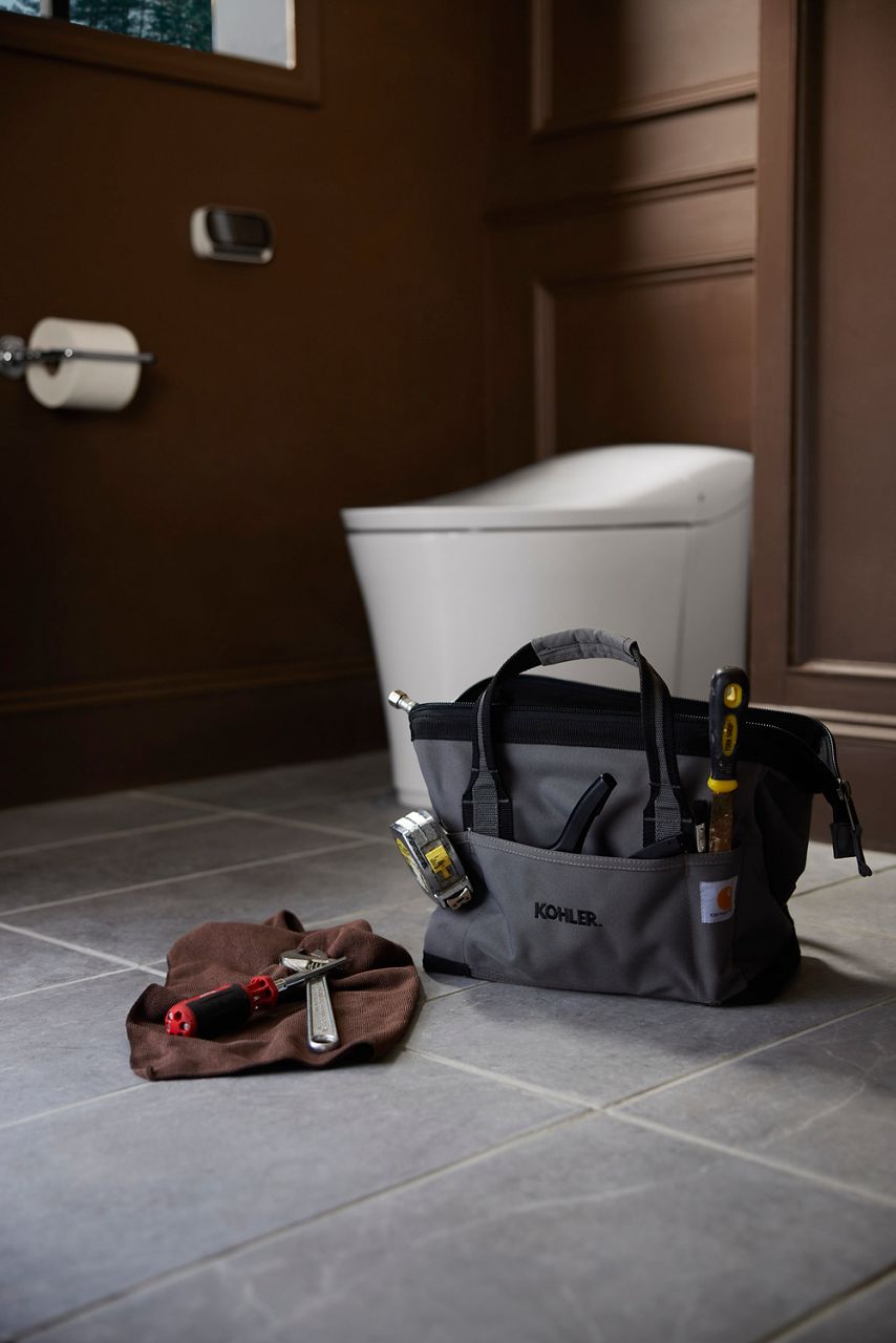 Tool bag and plumbing tools on bathroom floor with toilet in background for DIY toilet installation.