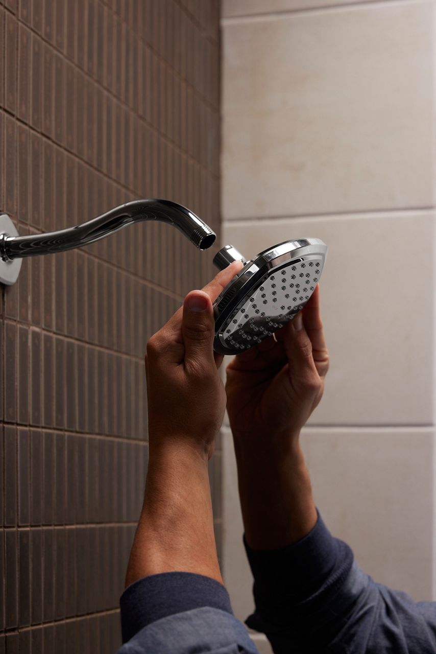 Close-up of hands tightening a shower head onto a shower arm in a tiled shower.