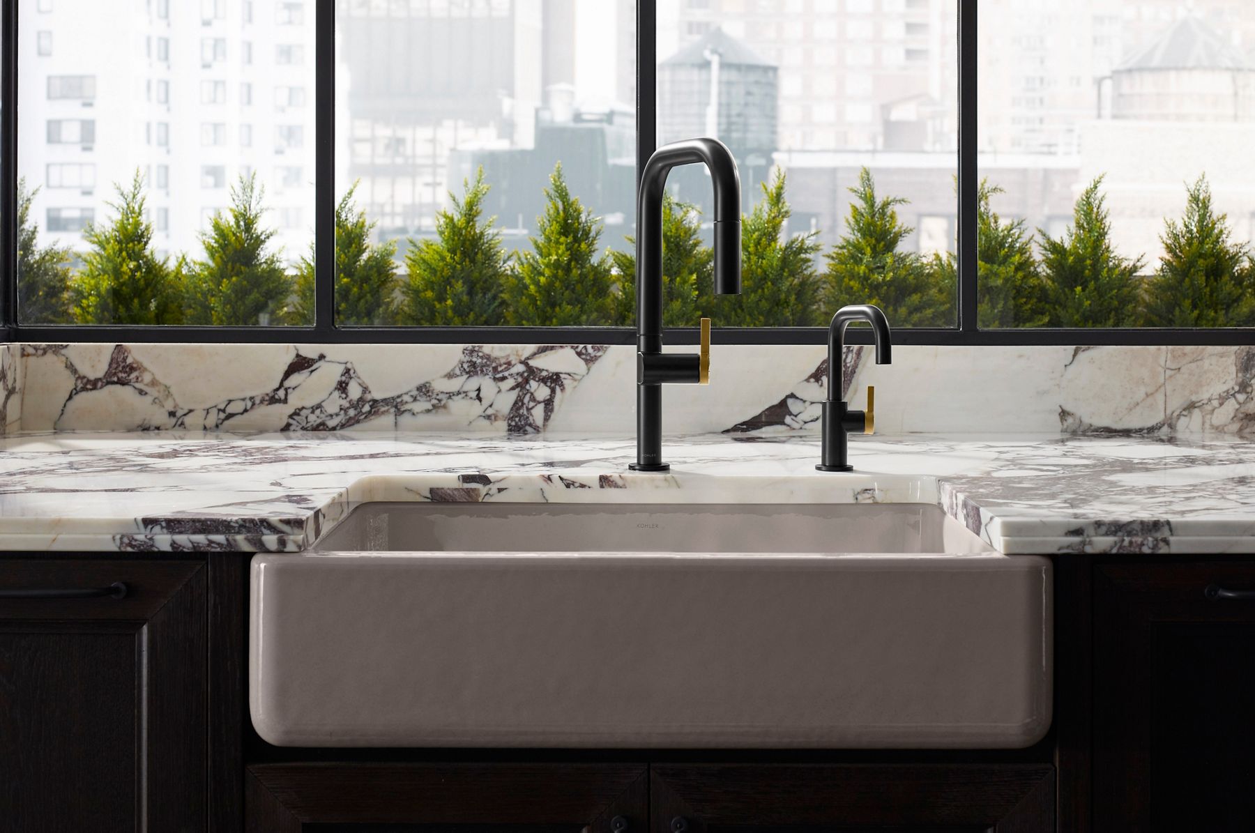 Modern kitchen featuring a pair of matte black faucets with sleek, minimalist designs. One faucet is taller with a high-arc spout, while the other is shorter, both with matching gold handles. The countertops are made of luxurious white marble with bold dark veining, and large windows in the background offer a view of greenery, adding a natural touch to the contemporary space.