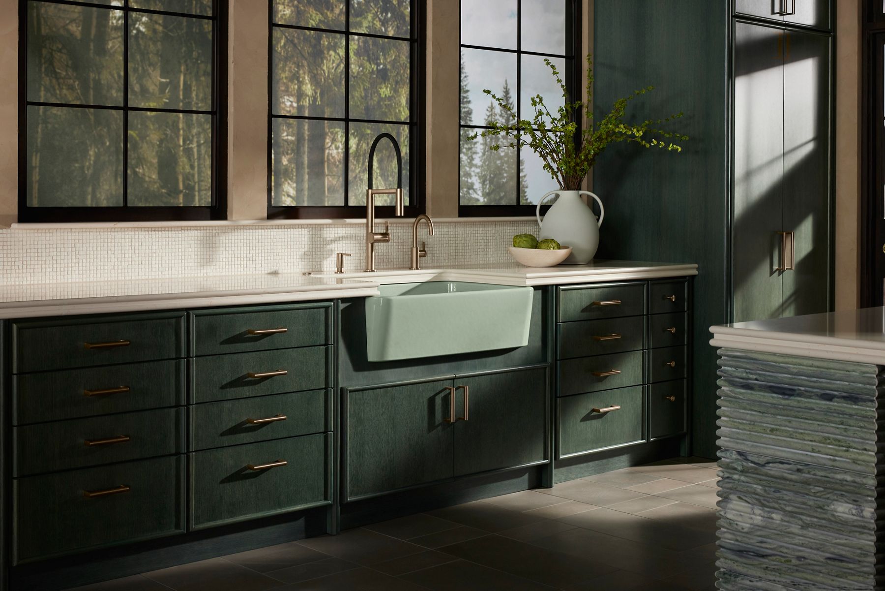 Modern kitchen with a farmhouse-style sink and a set of brushed nickel faucets, including a high-arc faucet, a smaller faucet, and a soap dispenser. The backsplash features small, white mosaic tiles, and large windows in the background offer a view of the greenery outside, adding natural light to the space.