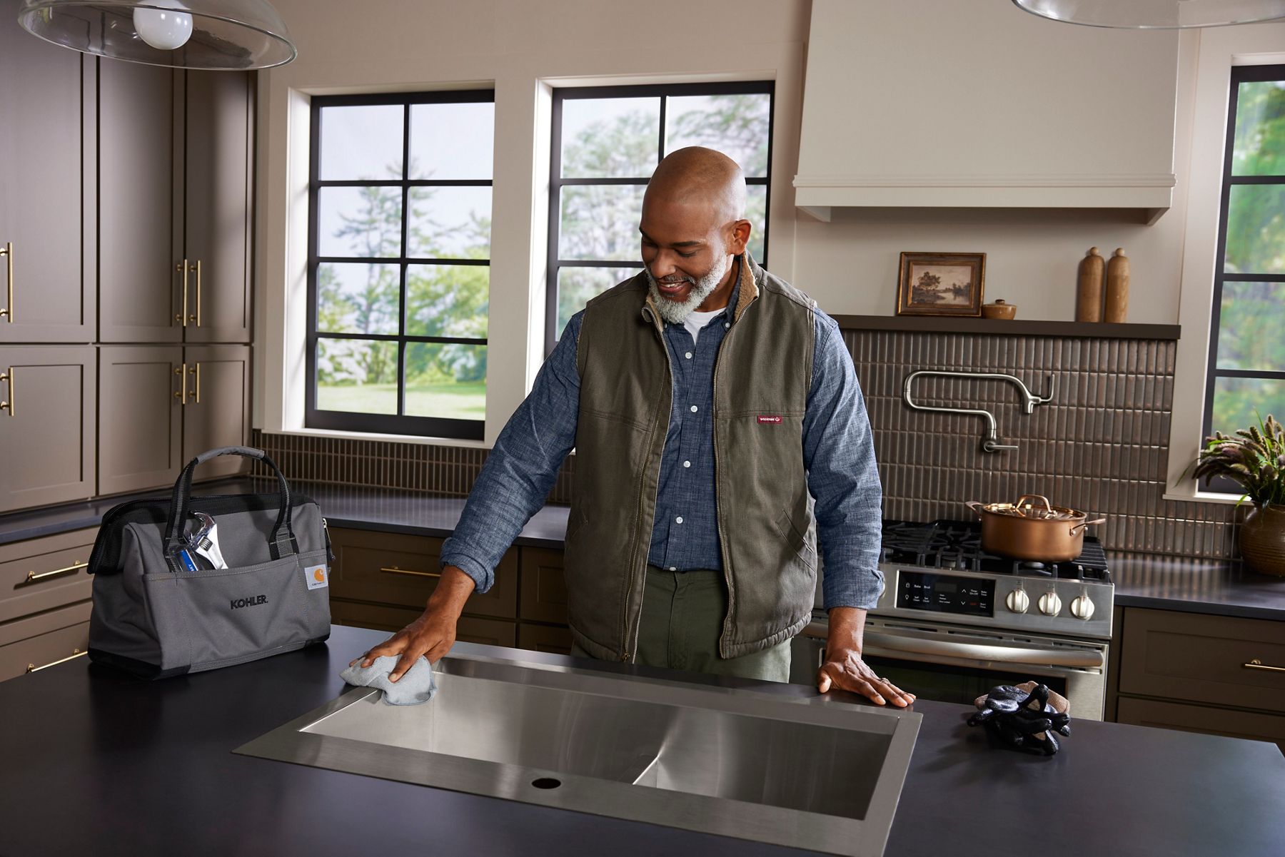 A person cleaning a kitchen sink beside a KOHLER tool bag on a counter in a modern kitchen.