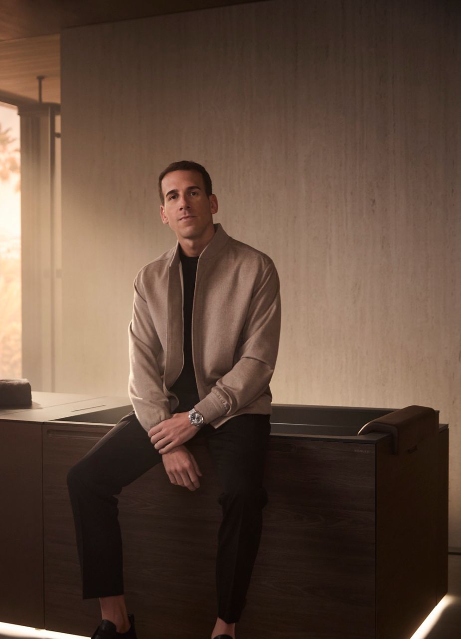Dr Jonathan Leary, fondateur de Remedy Place, assis sur le bord d’une baignoire pour bain de glace moderne avec des panneaux en bois foncé. Il est vêtu d’une veste beige clair et d’un pantalon noir, et regarde directement la caméra avec une expression calme. Le décor est une salle de bain minimaliste chaleureusement éclairée, créant une ambiance de luxe et de détente.