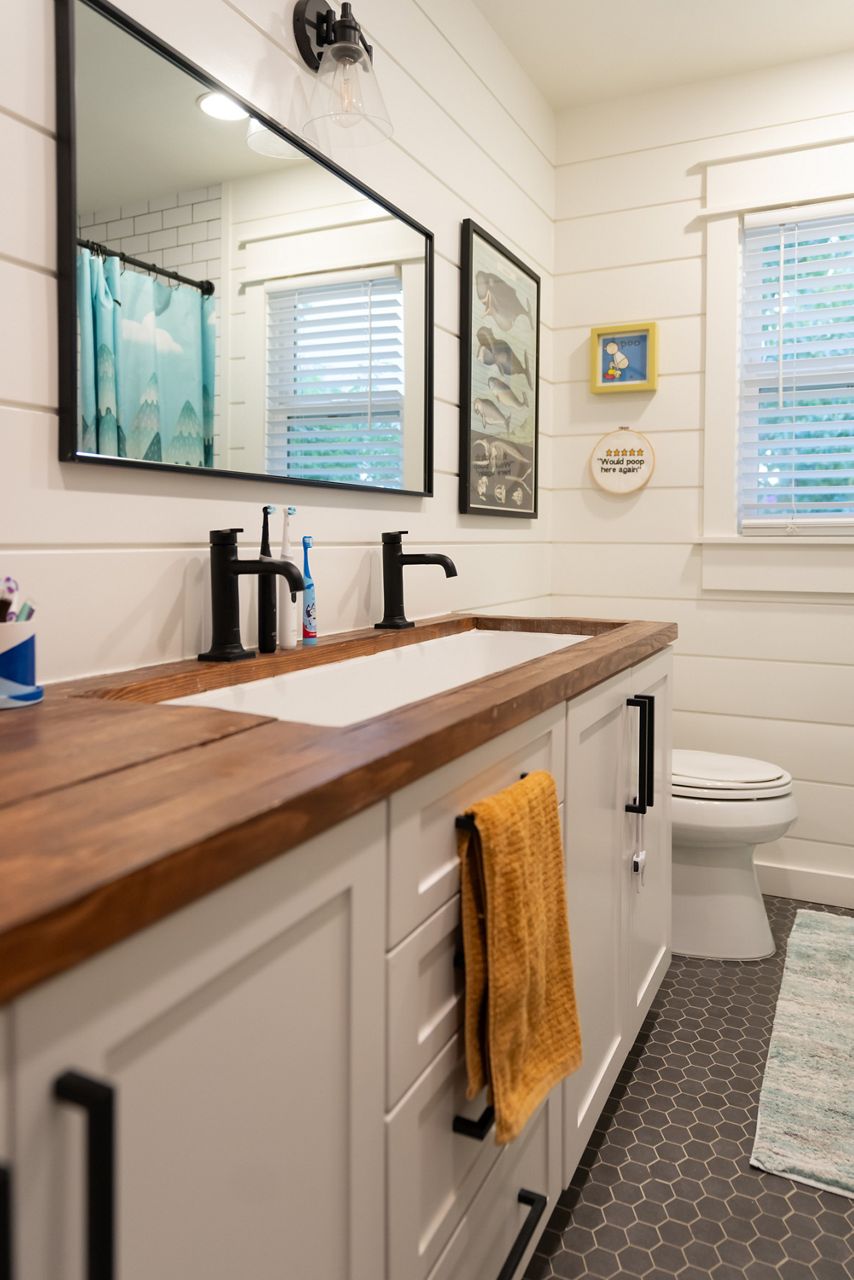 A modern bathroom with a wooden countertop, a single sink, black fixtures, a large rectangular mirror, and a toilet in a bright, white space with colorful decor.