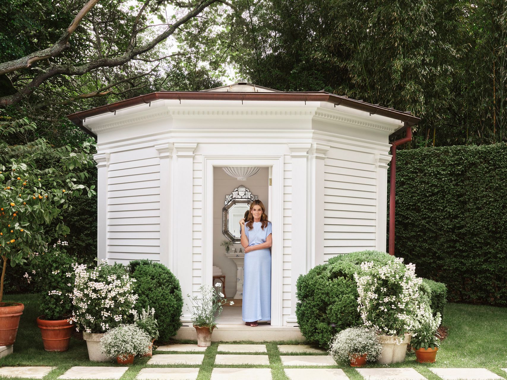 Aerin Lauder stands in the doorway of an elegant white garden pavilion surrounded by lush greenery and flowering plants.