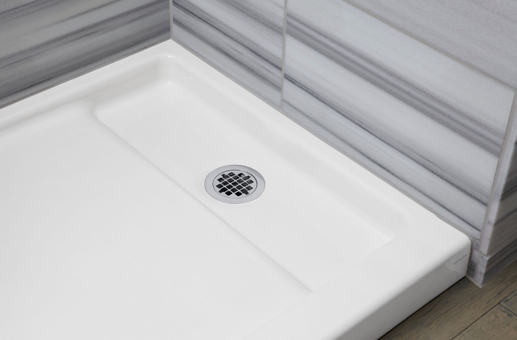 A close-up of a white enameled cast iron shower base and drain backed a gray wall