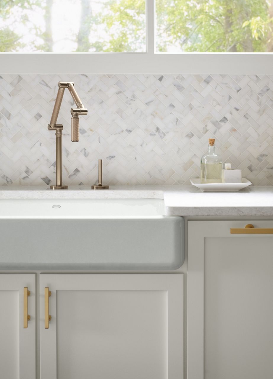 A bright sunlit kitchen features a KOHLER Whitehaven farmhouse kitchen sink and articulating kitchen sink faucet in front of Calacutta Borghini Mosaics marble tile
