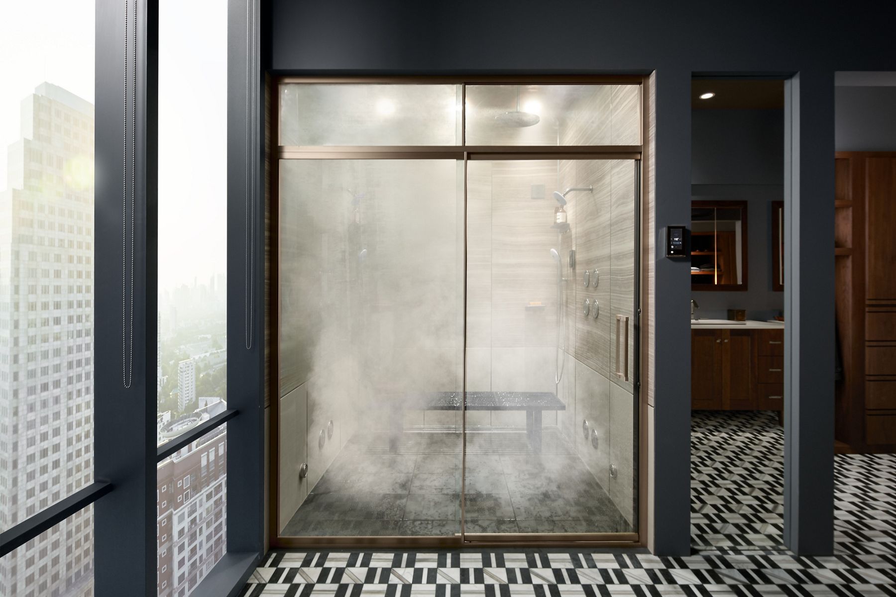 Steam fills a KOHLER steam shower with full glass doors with bronze trim in a dark gray bathroom with full length windows showing a city skyline.