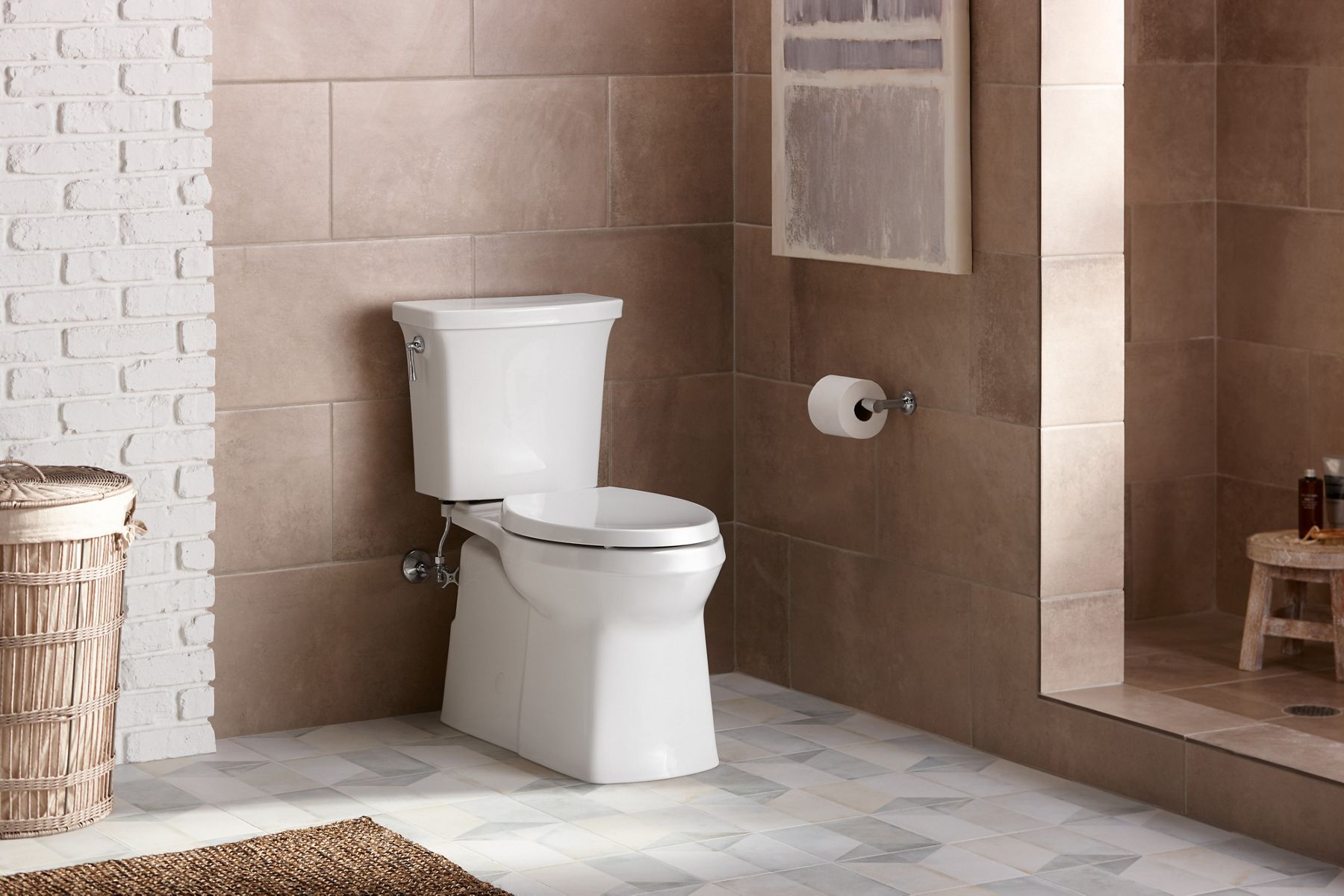 Une toilette allongée blanche Corbelle de KOHLER avec technologie de chasse d’eau à tourbillon Revolution 360 dans une salle de bain carrelée beige avec un grand panier à linge en osier.