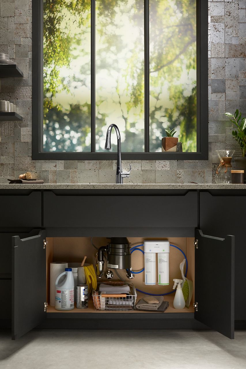 Modern kitchen with an open cabinet below the sink, revealing an organized storage space containing cleaning supplies, a water filtration system, and a garbage disposal unit. The countertop features a polished chrome faucet, and a large window above the sink offers a blurred view of greenery outside, adding a natural element to the sleek, contemporary design of the space.