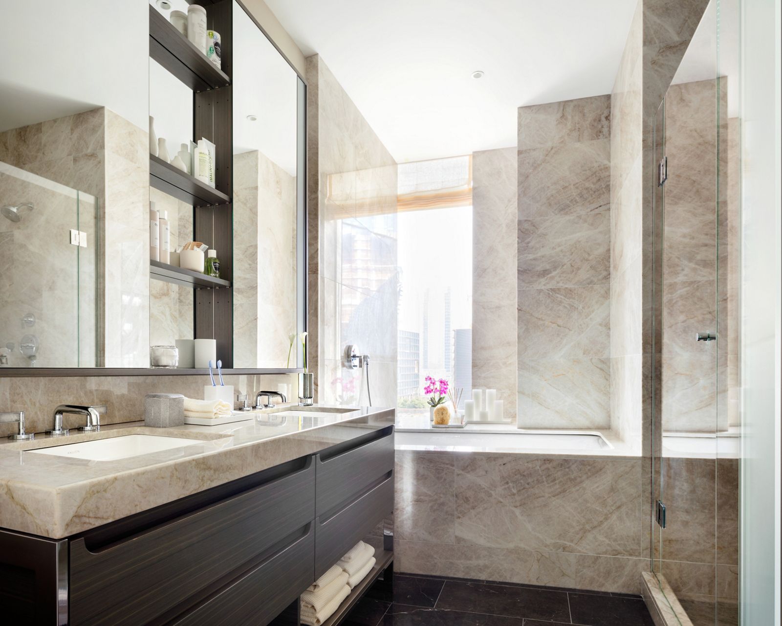 Interior view of a bathroom of One Hudson Yards featuring light brown marble on the walls and countertop with undermount sinks and bath under a large mirror reflecting the large glass-enclosed shower. 