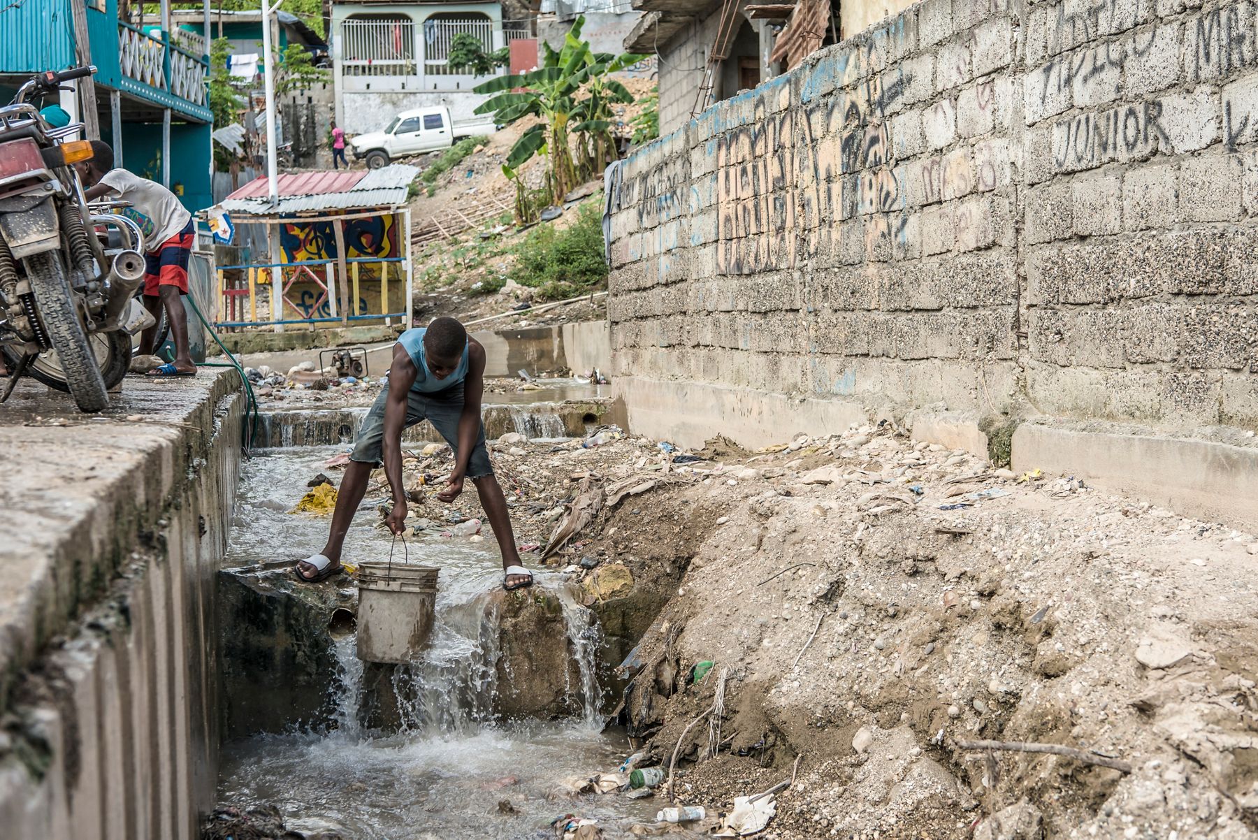 A man standing in a drainage canal collecting water with a bucket in an urban environment, surrounded by buildings, motorcycles, and a concrete wall.