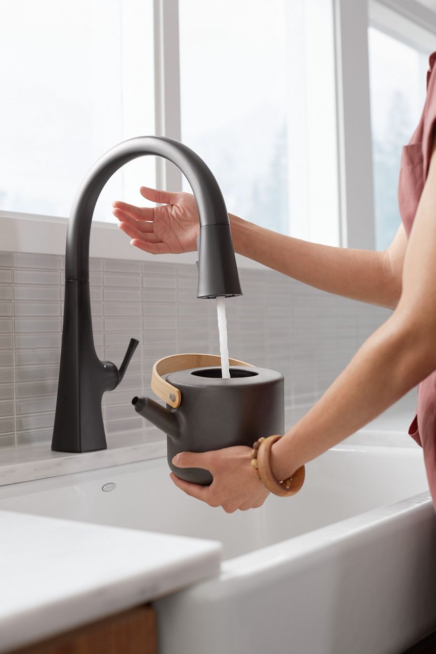 A person holding a black pitcher under a matte black faucet and using her right hand to turn the faucet on using its touchless sensor.