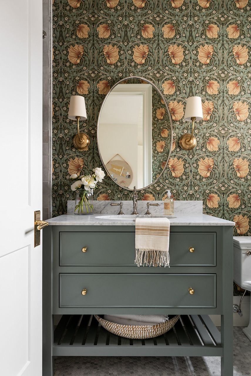 A bathroom vanity in dark pale green with matching patterned floral wallaper and an oval mirror with sconce lights.