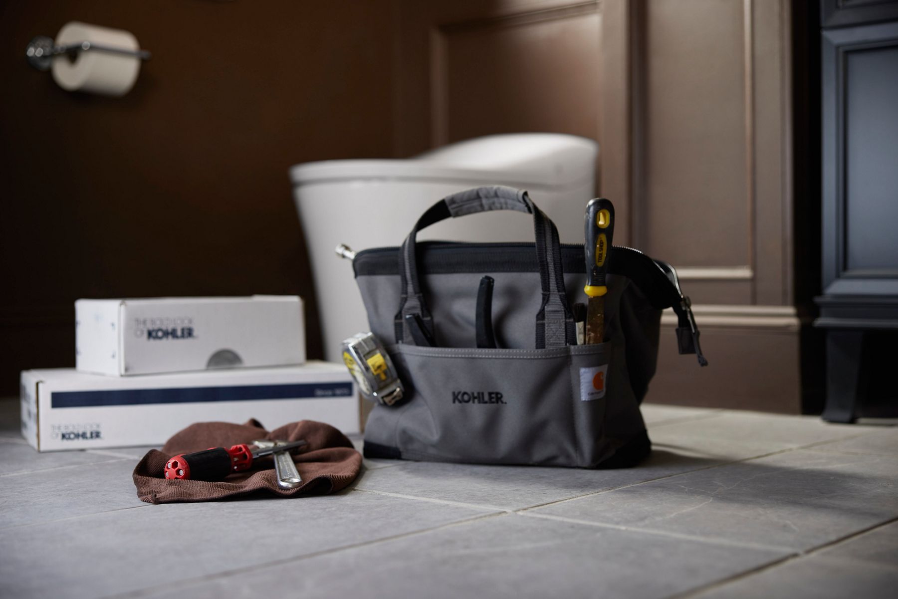 A bathroom setting featuring a Kohler-branded tool bag placed on the floor, filled with various tools such as a screwdriver and measuring tape. Nearby are boxed Kohler products, a cloth with a tool on top, and a toilet in the background. The scene suggests a plumbing or bathroom installation or repair project.