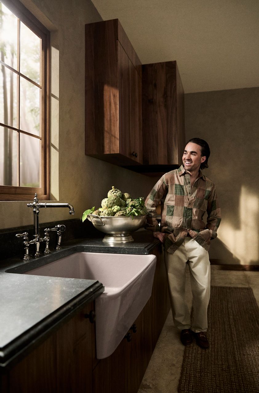 Jake Arnold standing next to a white farmhouse-style kitchen sink with a tall chrome faucet and a bowl of artichokes on the counter.
