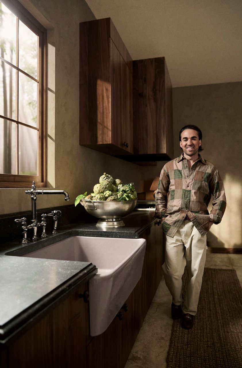 Jake Arnold standing next to a white farmhouse-style kitchen sink with a tall chrome faucet and a bowl of artichokes on the counter.