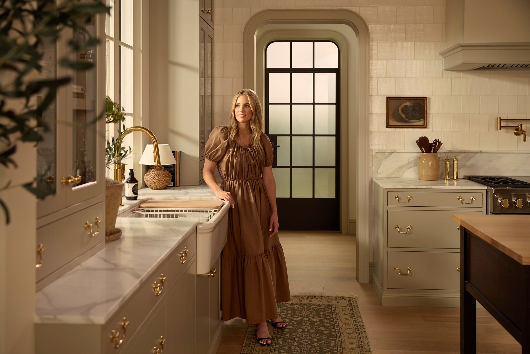 Shea McGee wearing a flowing khaki dress standing in a warmly lit kitchen that has gold hardware, neutral stone countertops, and wood accents.
