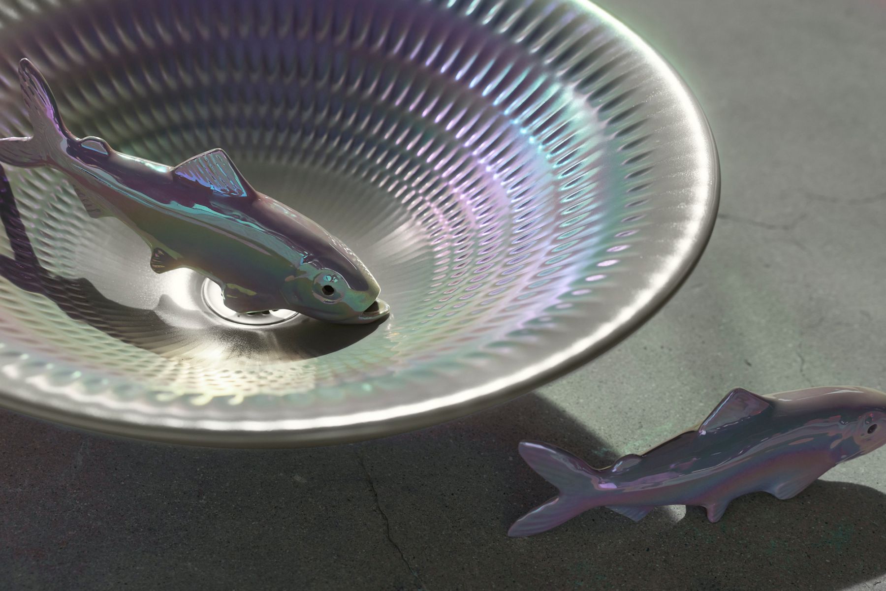 Close-up view of a pearlized Kohler Derring sink from the Underlight installation, featuring a sculptural fish resting inside the iridescent basin with another fish visible nearby on a concrete surface