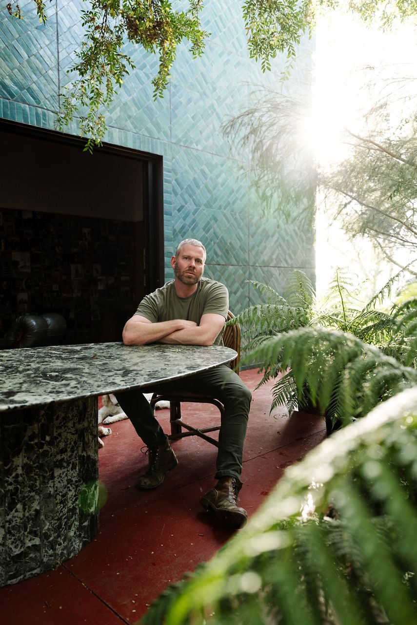 Richard Christiansen, fondateur de Flamingo Estate, vêtu d’une tenue entièrement verte, est assis à une table d’extérieur en marbre, entouré de plantes luxuriantes dans un jardin paisible.