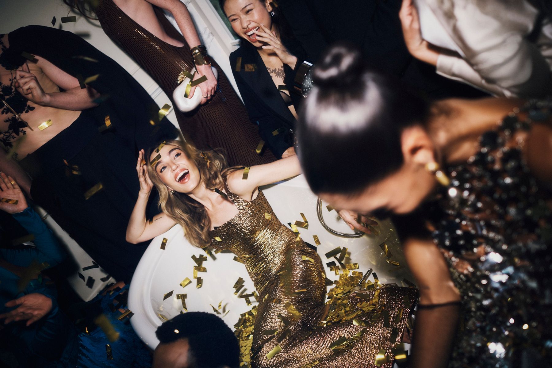 A person dressed in a gold gown sitting in a bathtub without water and glitter in the air to create a festive party scene with other people in the foreground.