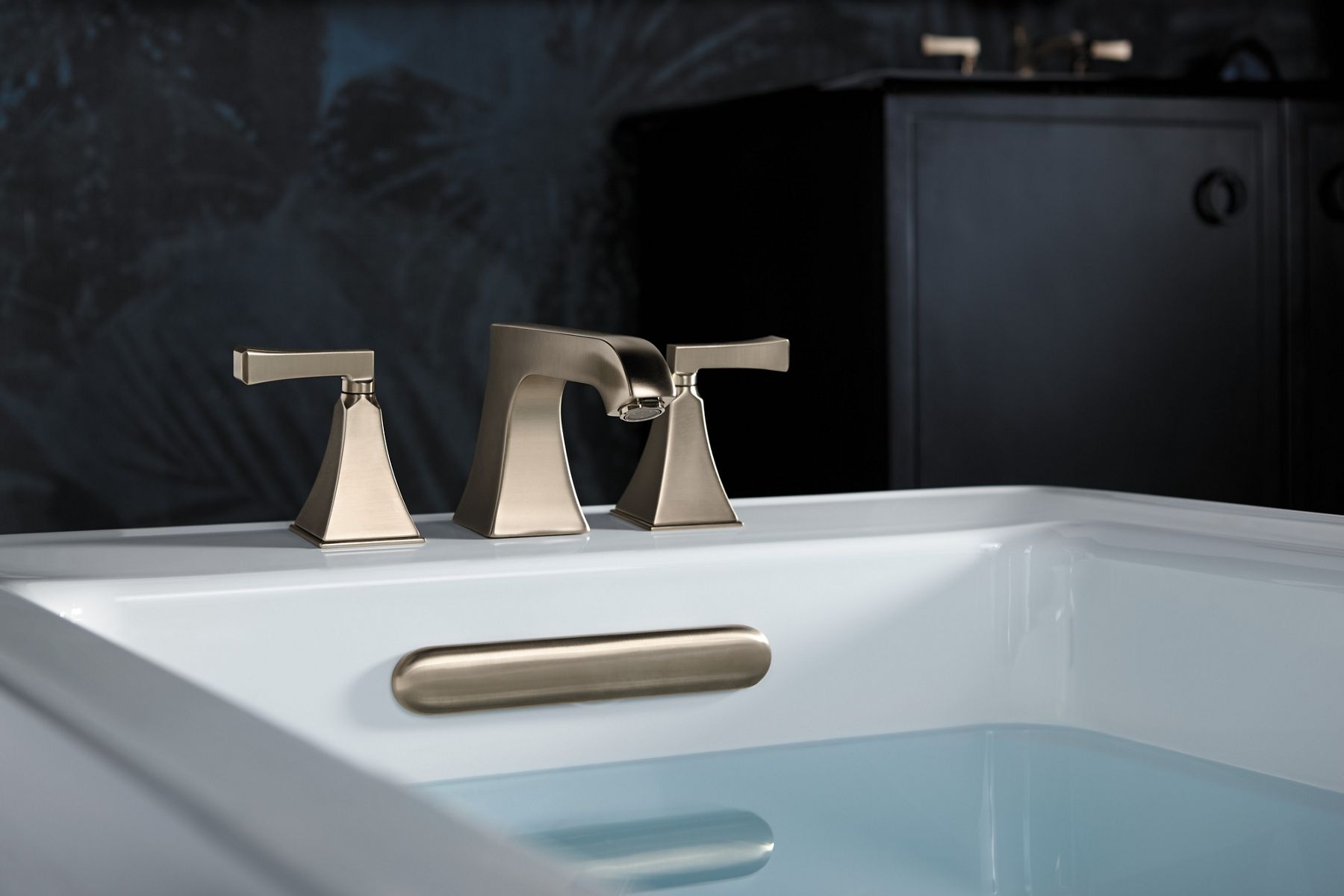 A brushed bronze bath faucet and overflow drain in a white bath filled with water.