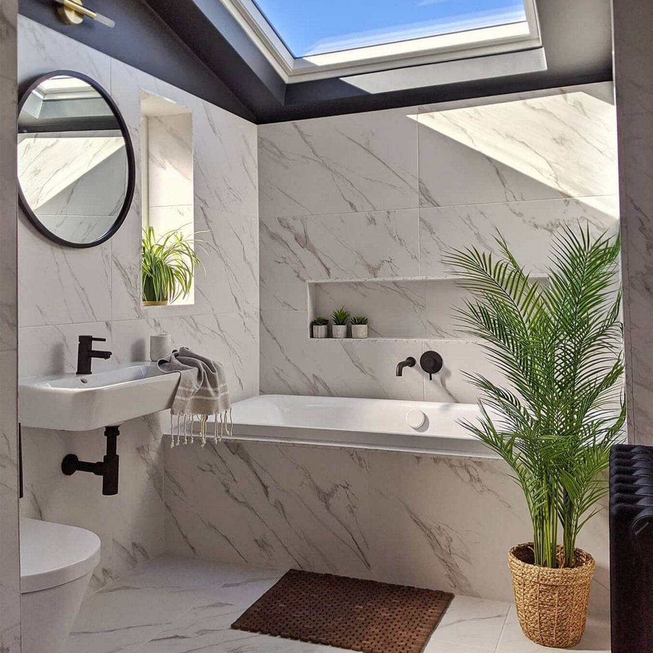 An after picture of @thehalfdonehouse's renovated bathroom with marble style tiles, a skylight window, black fixtures and a large plant.