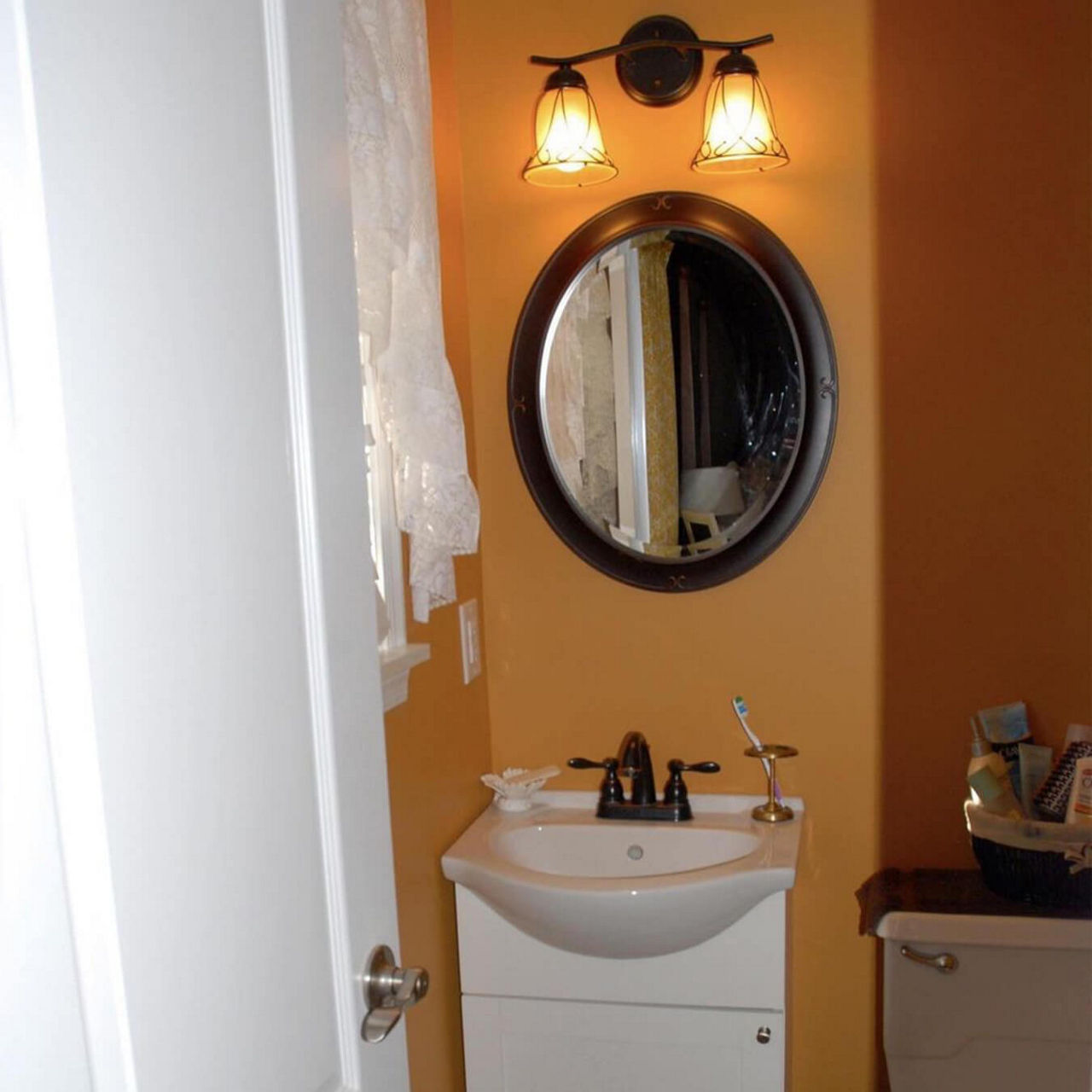 A before picture of @thistle.harvest's bathroom with toilet and a small sink against an orange wall, and an oval mirror and vintage style sconces above them.