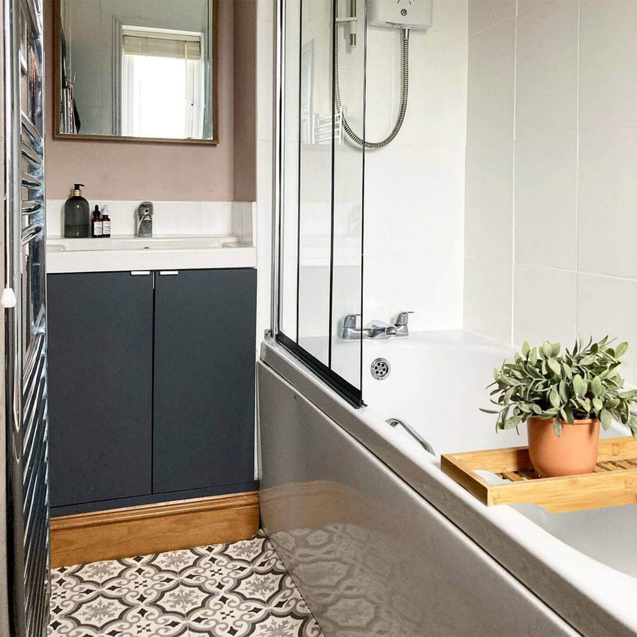 An after picture of @katieelvins's renovated bathroom with dark grey under-sink cupboards, geometric vinyl flooring and a shower-over bath.