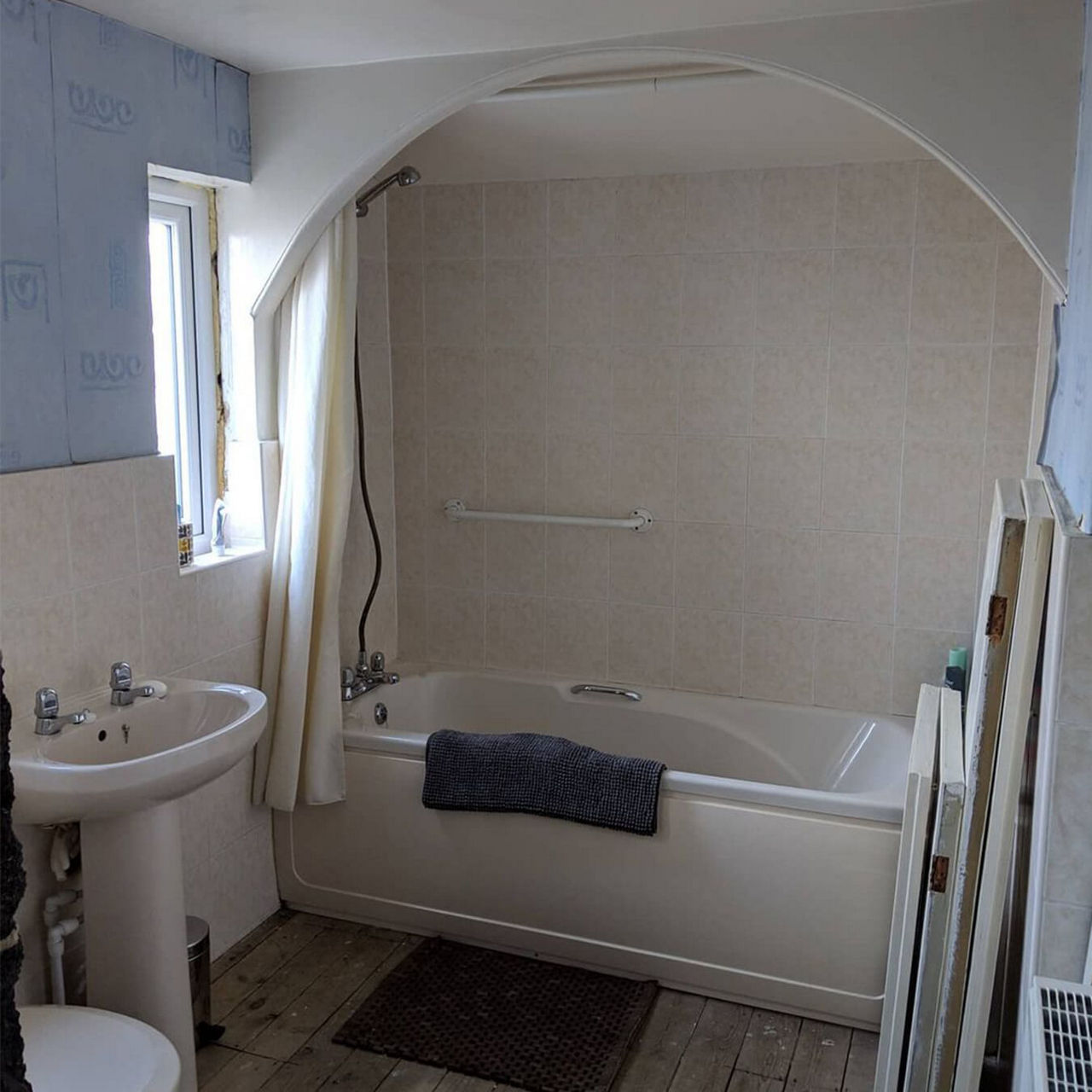 A before picture of @thehalfdonehouse bathroom with a wood floor, shower-over-bath with an archway and a window.