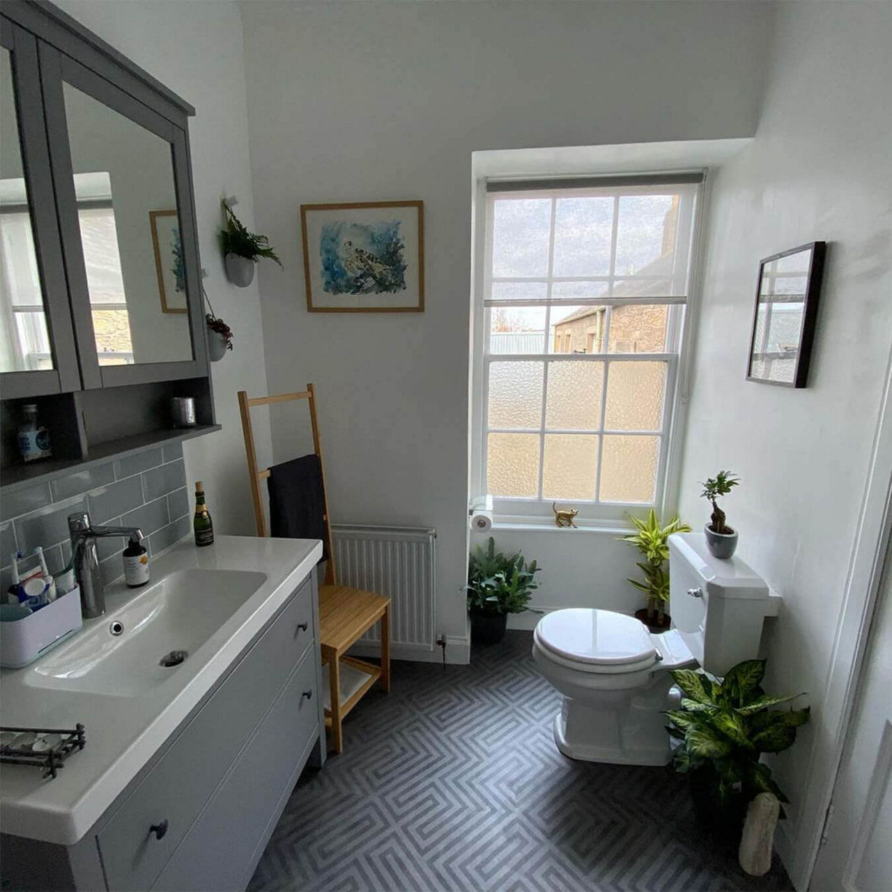 An after picture of @ailsa.bm's renovated bathroom with grey geometric flooring, a grey and white sink vanity with grey overhead cupboards, a toilet and many plants.