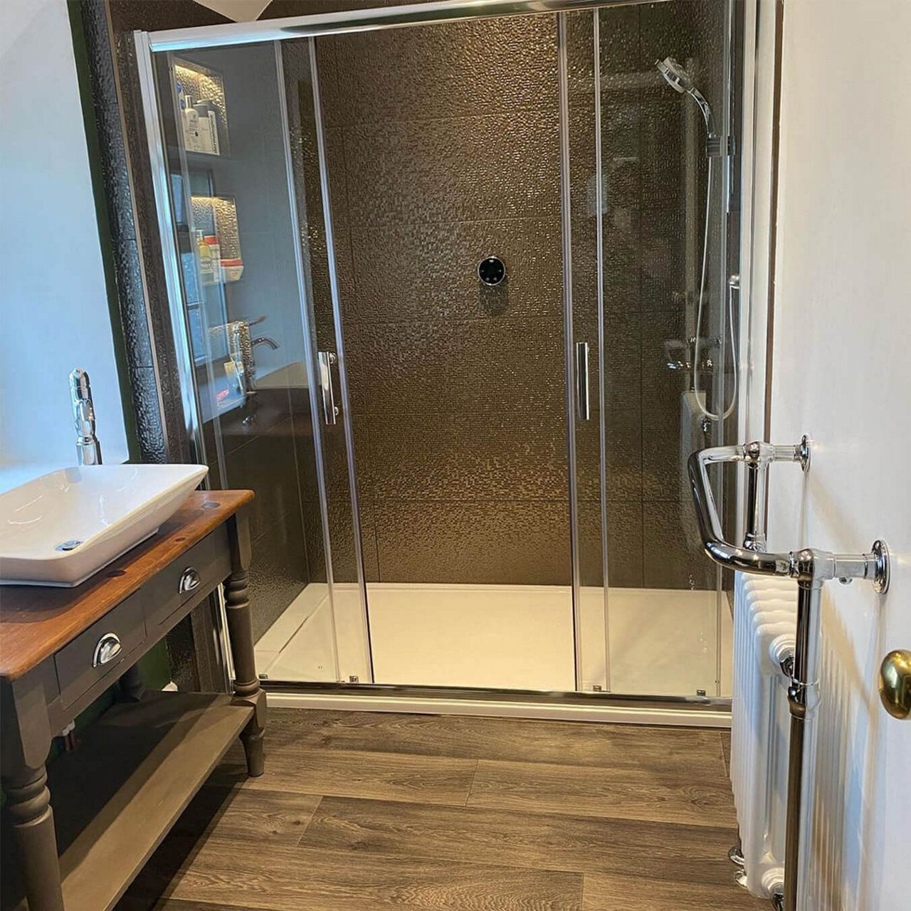 An after picture of @compton_randle's renovated bathroom with wood floors and a large shower enclosure with dual sliding doors and dark green tiles.