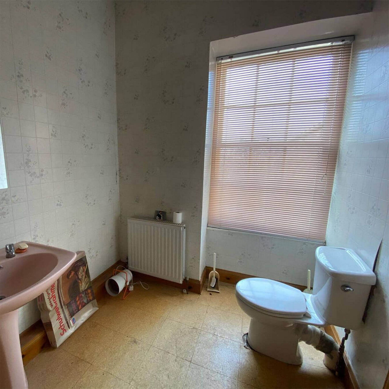 A before picture of @ailsa.bm's bathroom with brown vinyl flooring, floral wall tiles, a toilet and large window.