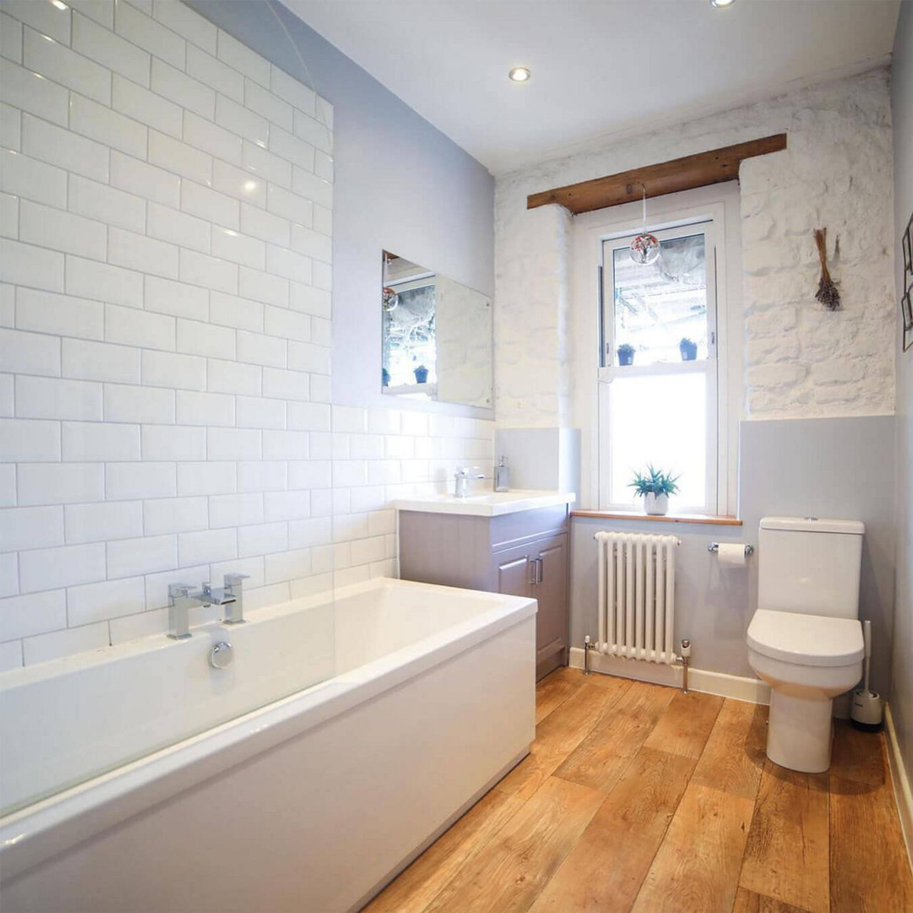 An after picture of @restorationcouple's renovated bathroom with wooden floors, white and grey walls and vanity, a corner bathtub and a toilet.