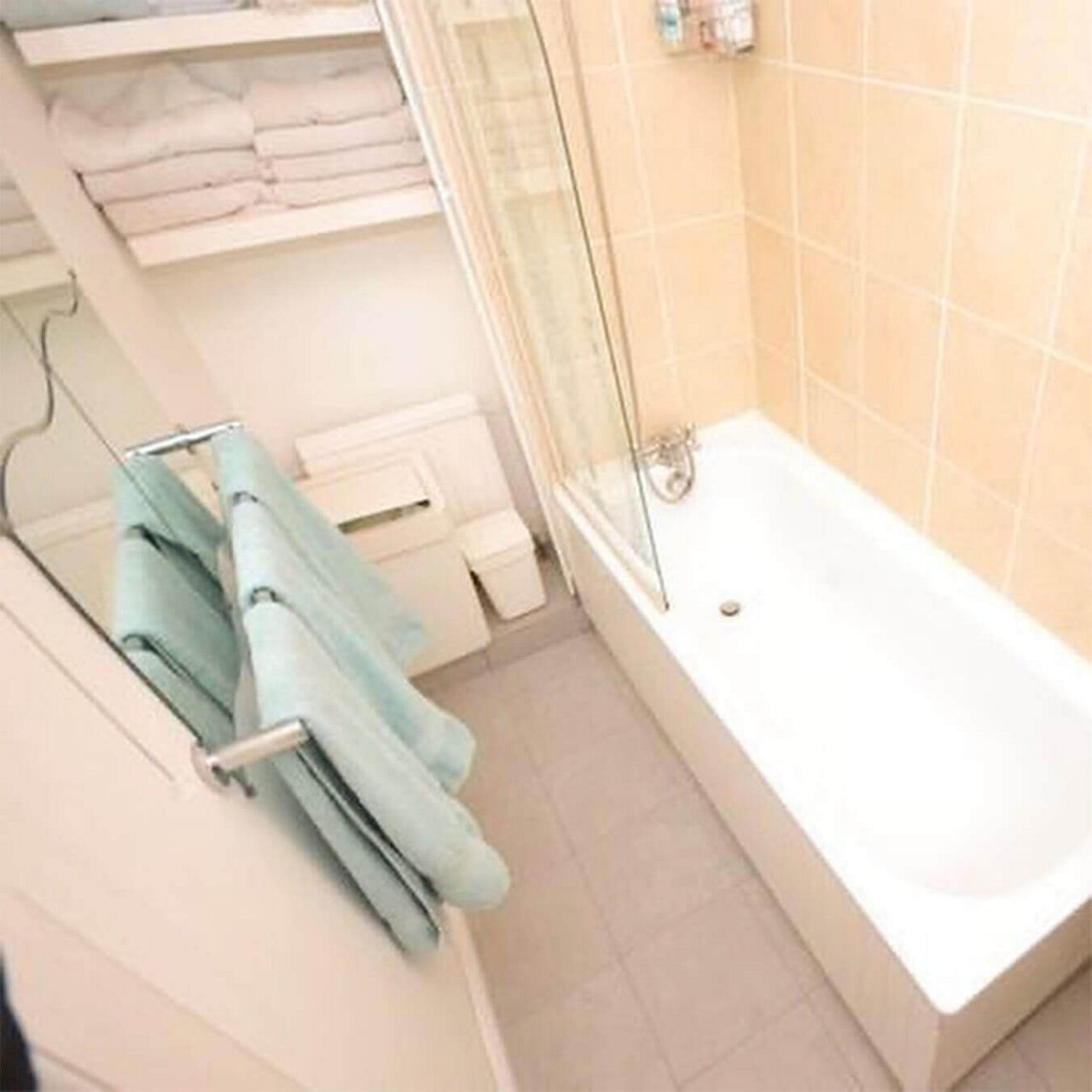 A before picture of @katieelvins's bathroom with a shower-over-bath, beige tiles and white shelves with towels.