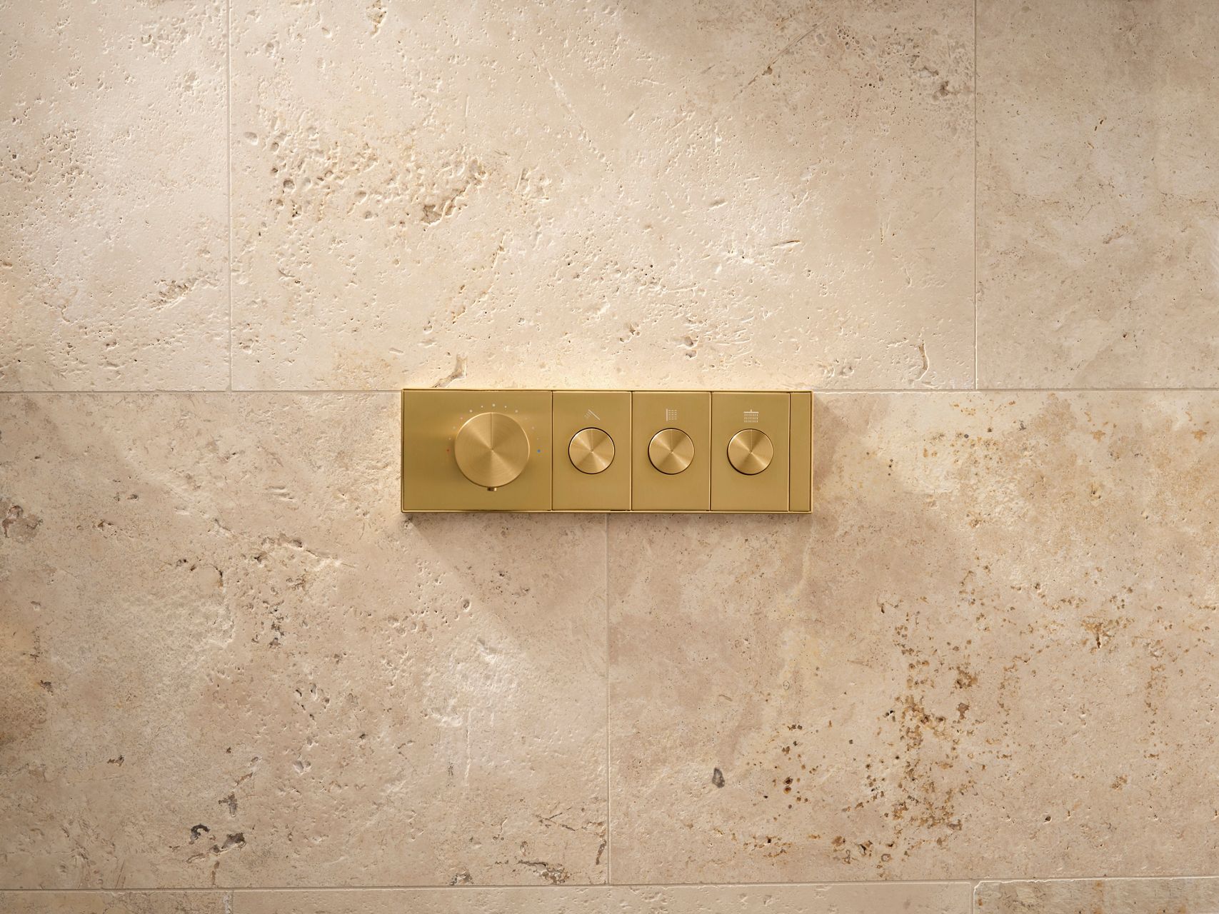 A brushed brass mechanical shower control on a tan stone wall.