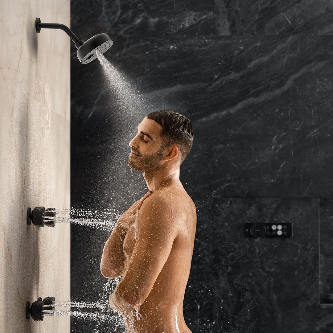 A man standing in a shower, enjoying water spraying from multiple side jets and an overhead showerhead. He is smiling with his eyes closed, one hand on his chest, with a black, textured wall in the background.