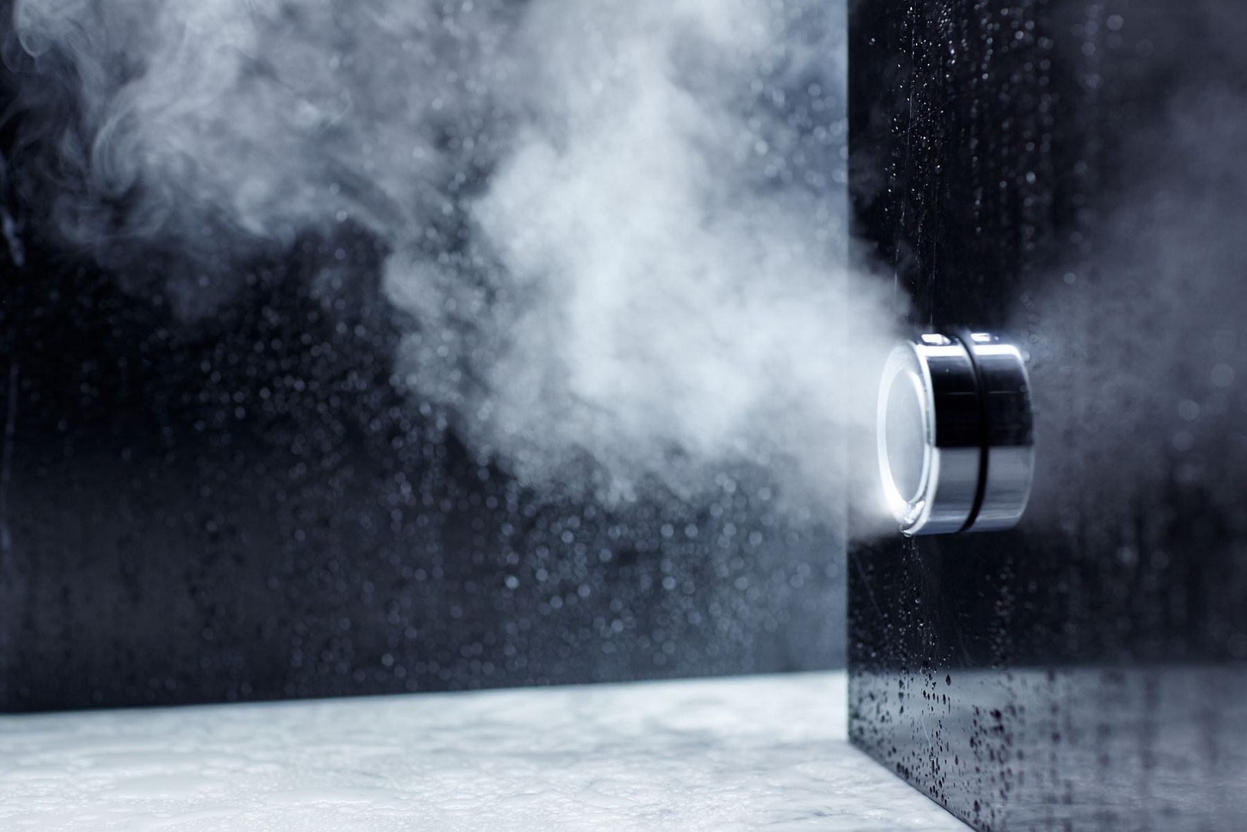 Close-up of a sleek, modern shower knob on a dark, wet surface with steam emitting from around the knob, creating a misty atmosphere.