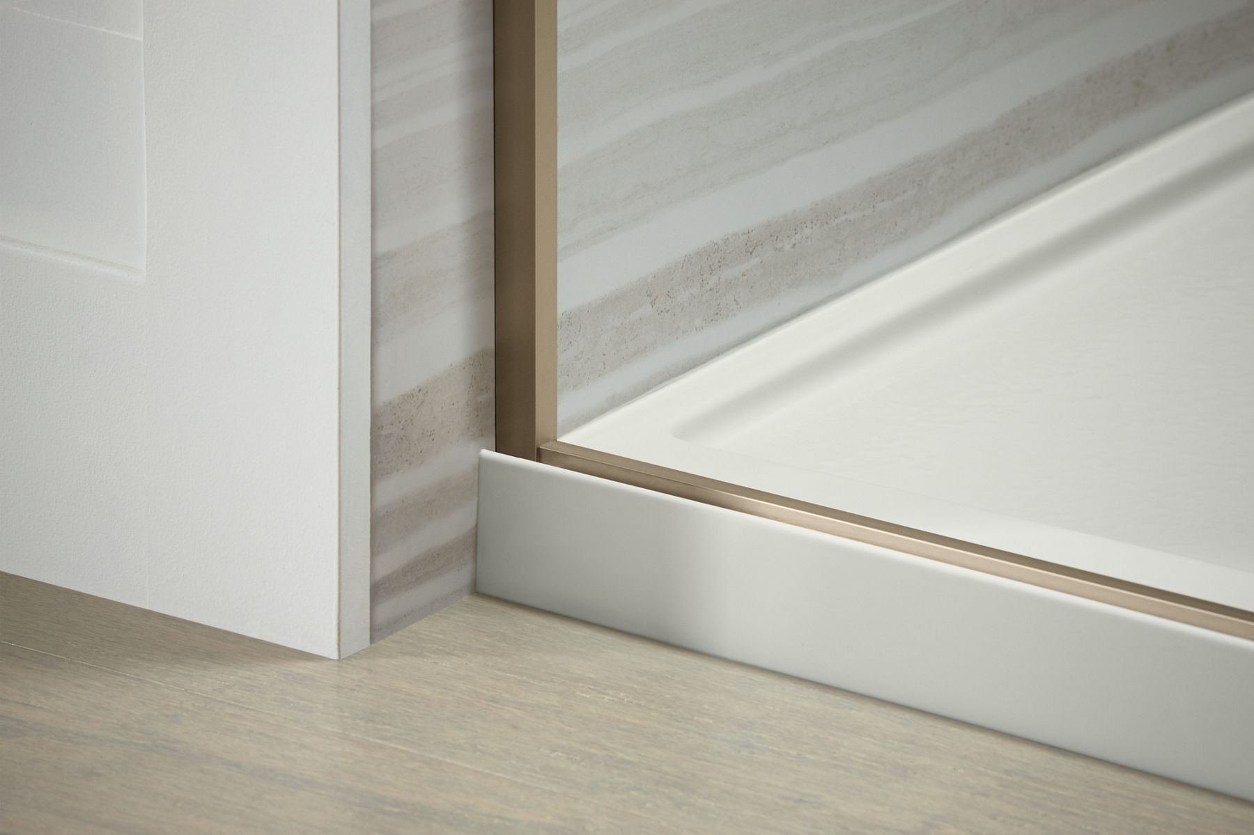 Close-up of the corner of a glass shower enclosure with a gold frame, showing the intersection of the glass, frame, and tiled wall, along with a white shower base and wooden floor.