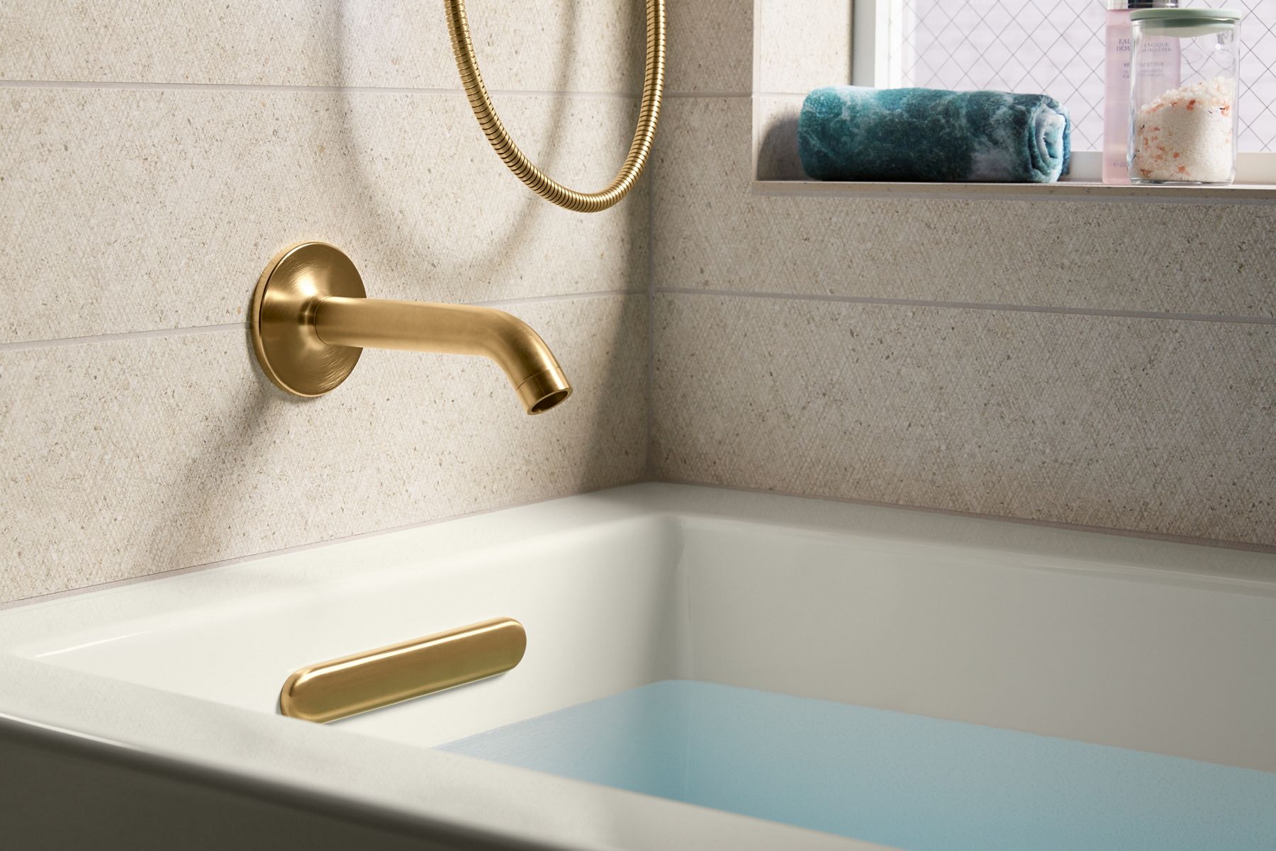 Close-up of a gold-colored bathtub spout with a matching handle mounted on a beige tiled wall. The spout is filling a white bathtub with water, and a gold shower hose hangs nearby.