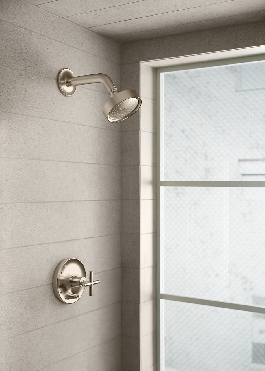 Close-up of a modern, brushed nickel shower handle with a cross-shaped design, mounted on a light-colored tiled wall.