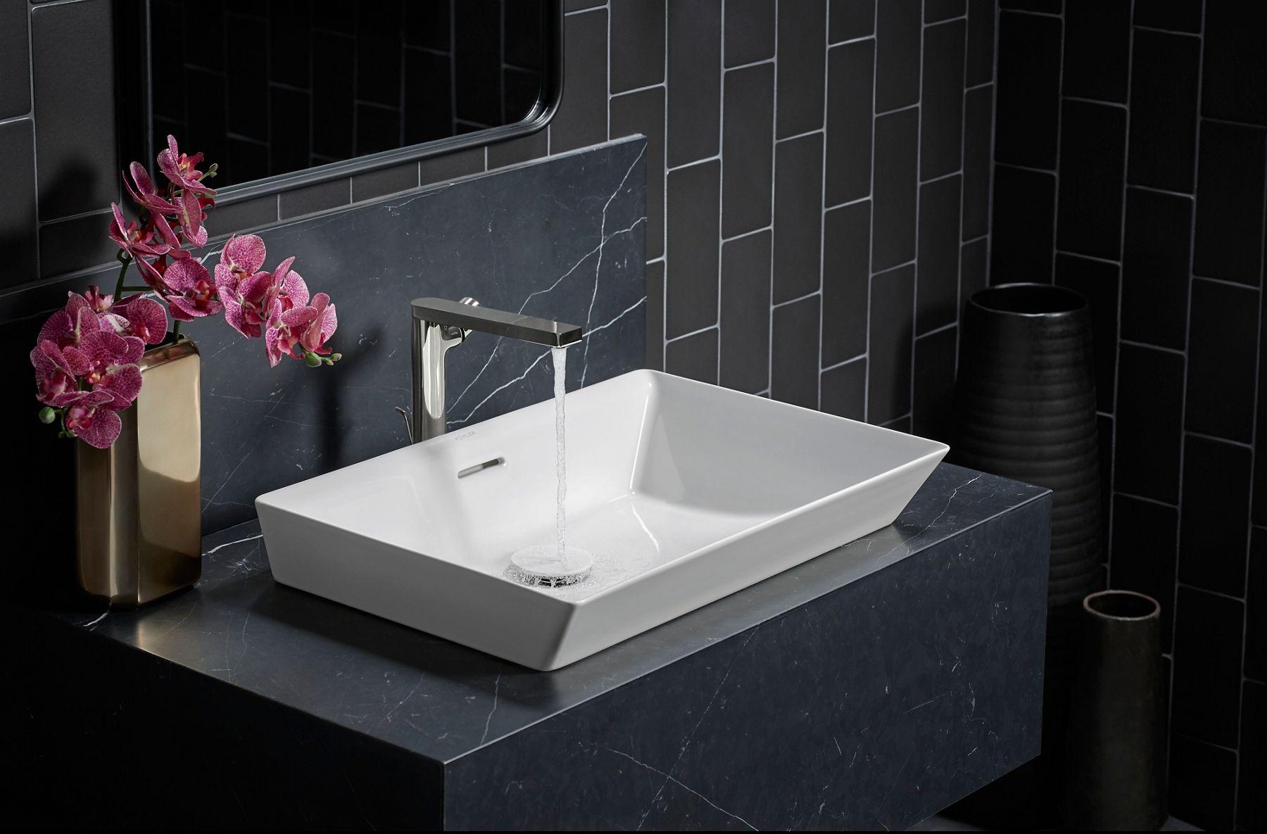 A close-up of a chrome bathroom sink faucet with running water over a a white virteous China bathroom sink on a black counter, backed by a black tile wall